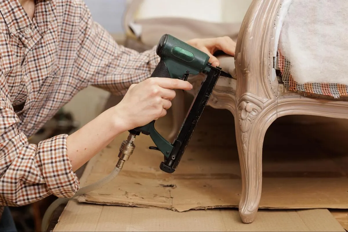 Upholstery technician using a staple gun to reupholster a wooden-framed armchair on a workshop bench